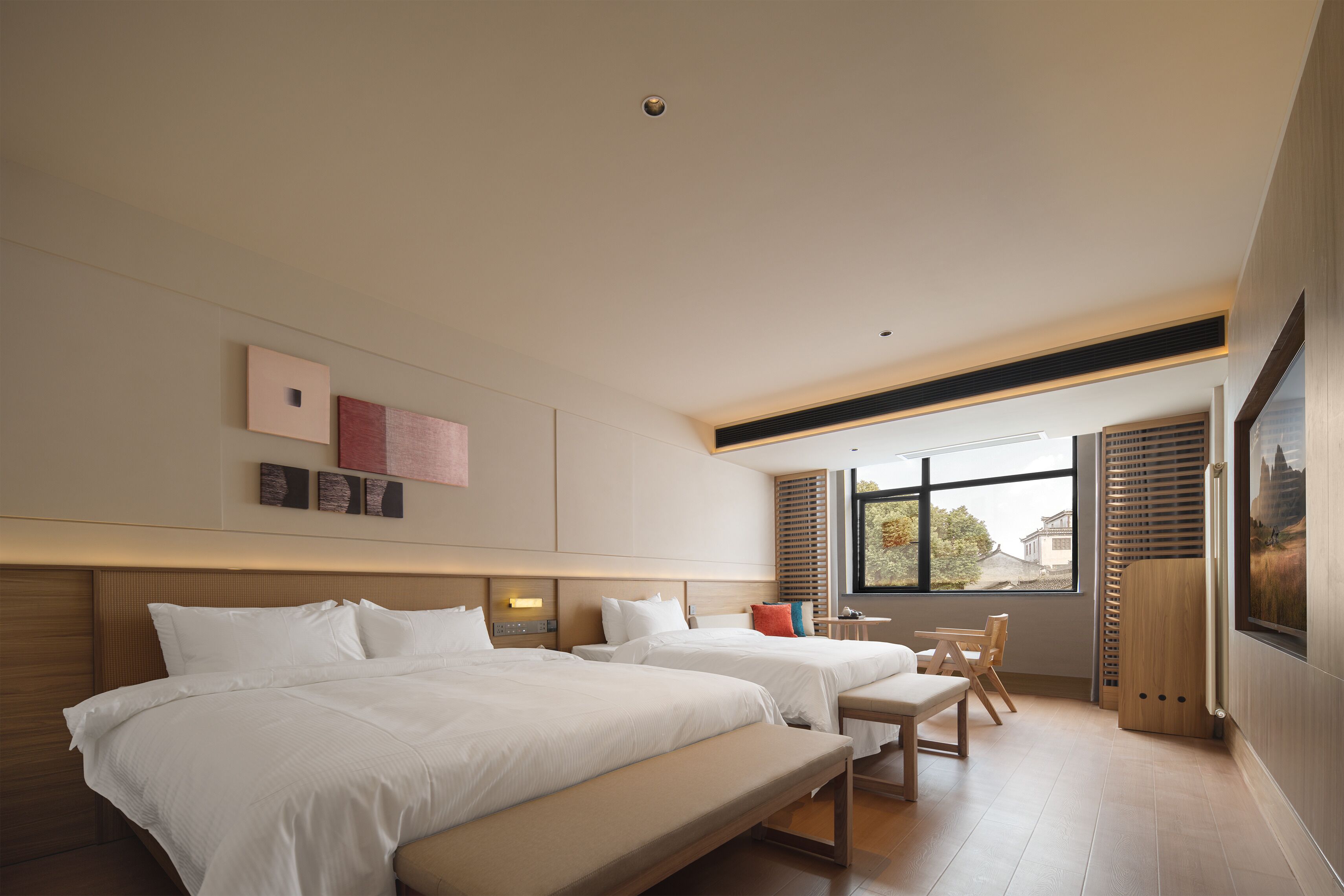 A modern hotel room with two beds, a bench, a small table, and a large window offering a view of the outside.