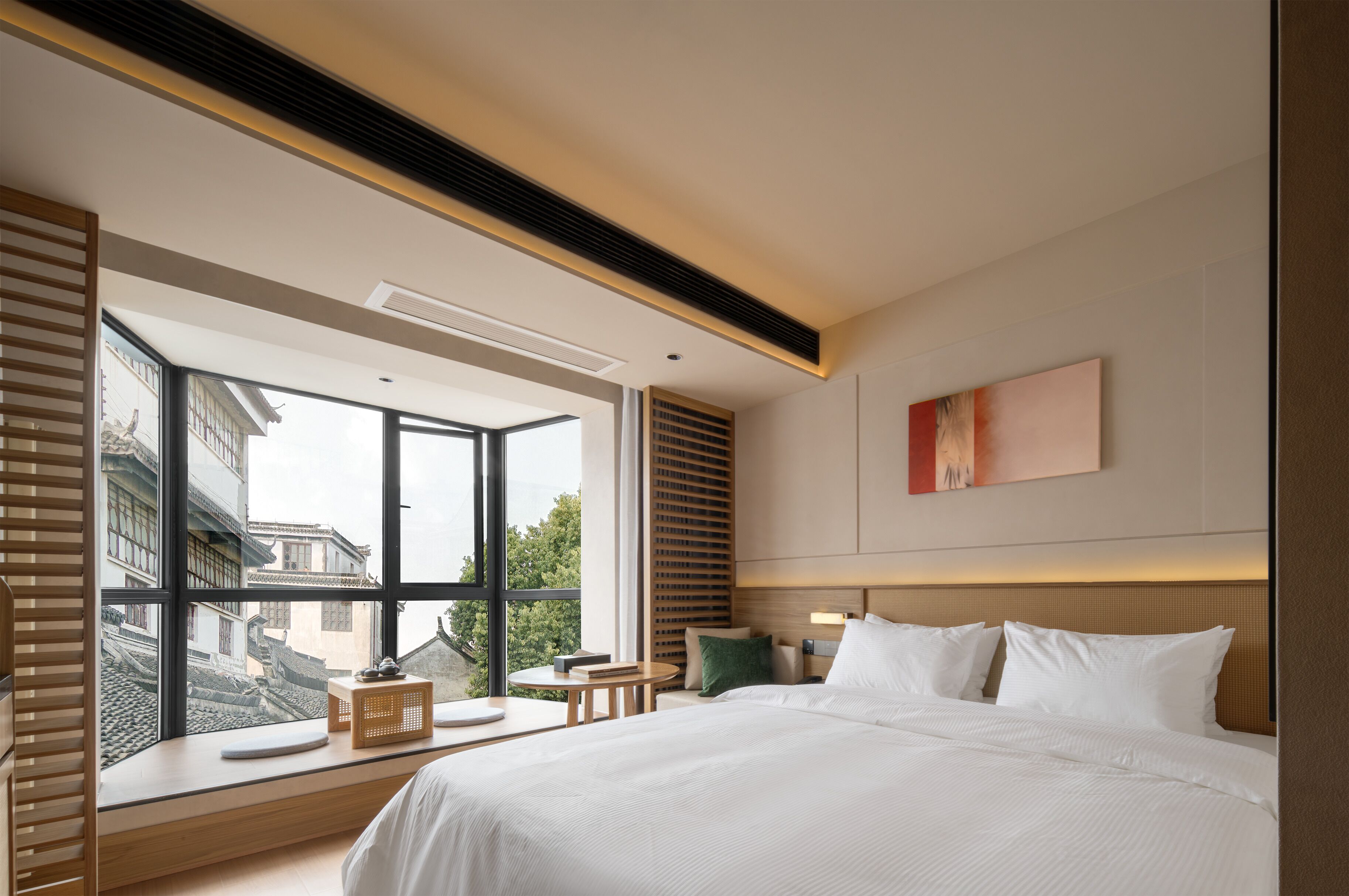 A modern hotel room with a large window, a bed, bedside tables, and a view of traditional buildings outside.