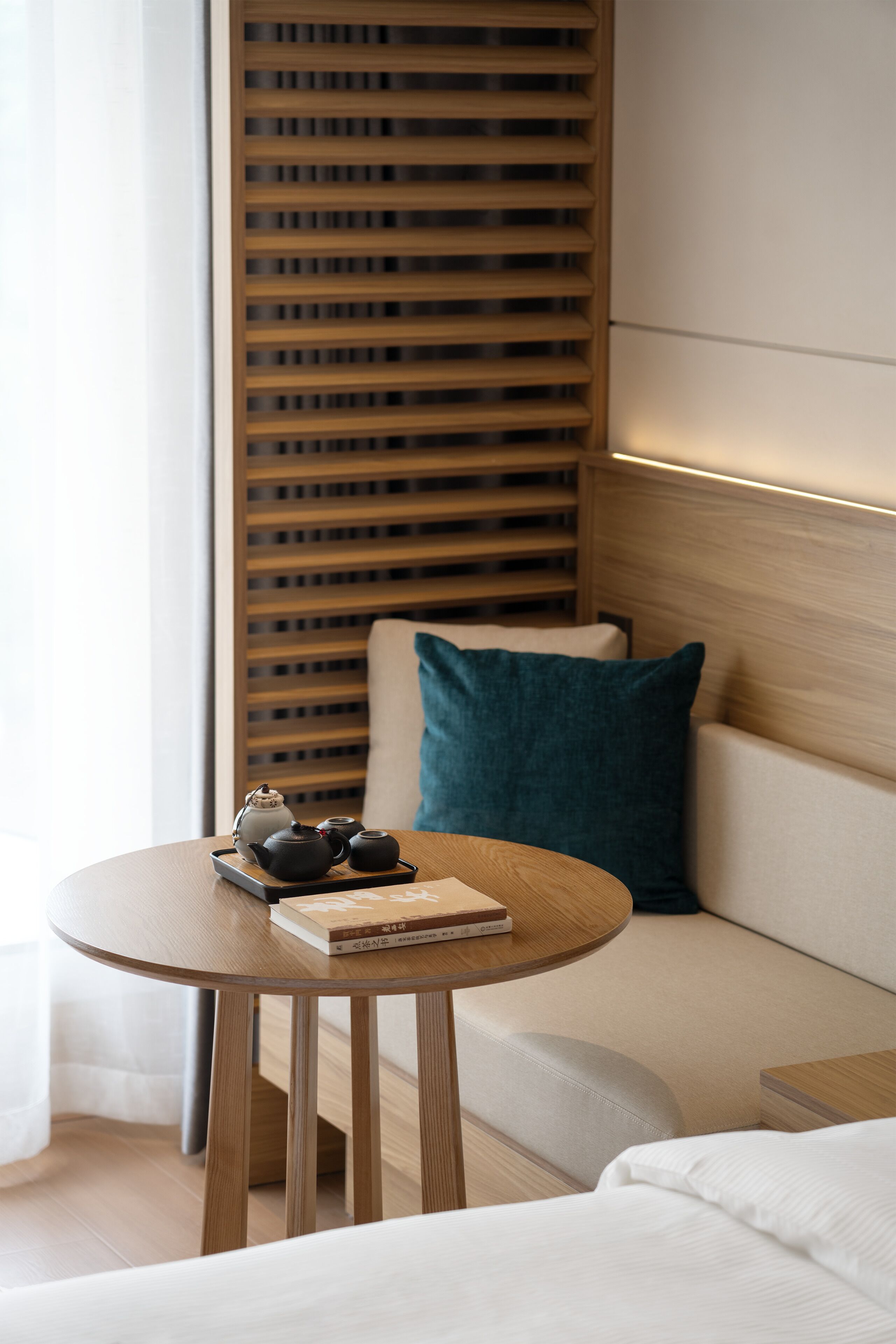 Quarto de hotel moderno com parede de ripas de madeira, mesa redonda de madeira com chaleira e livro, almofada verde-água e sofá branco.