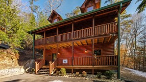 A two-story log cabin with a balcony, surrounded by trees and a stone wall.