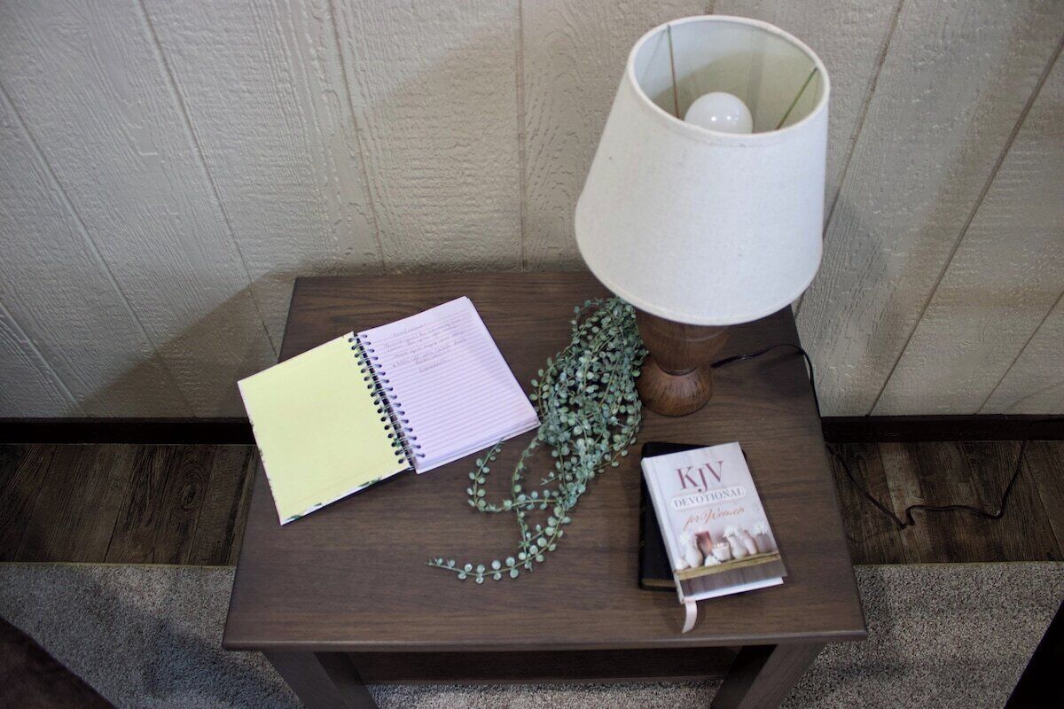 Ein kleiner Holztisch mit einem Spiralheft, einer Zierpflanze, einem Buch mit dem Titel 'KJV Devotional' und einer weißen Tischlampe.