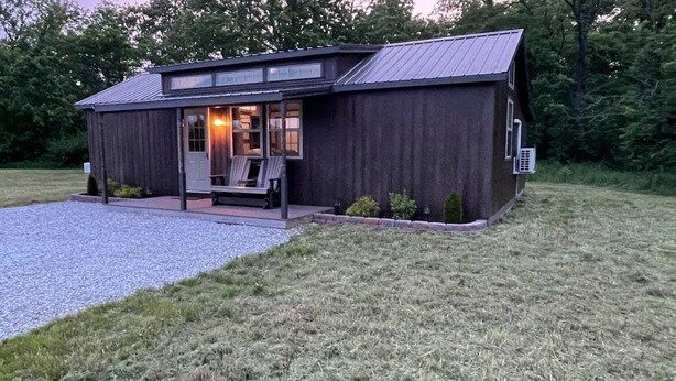 Een klein, rustiek gebouw met een metalen dak, een veranda met twee stoelen en een grindpad.
