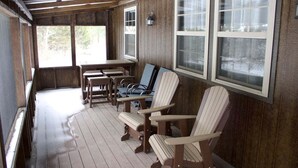 Een overdekt terras met houten vloer, Adirondack-stoelen, een tafel en grote ramen.