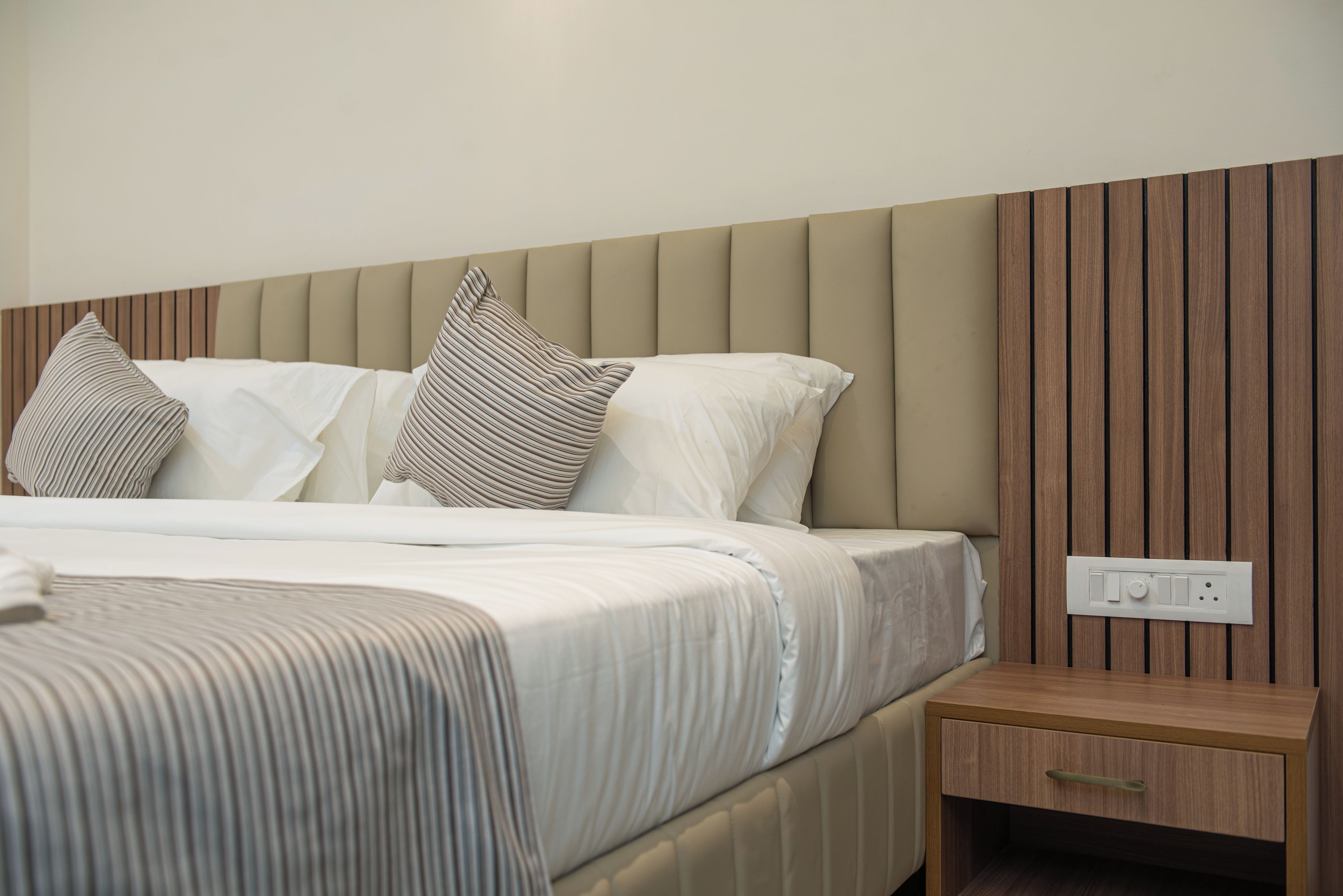 A neatly made bed with white linens and striped pillows, a wooden headboard, and a bedside table with a drawer.