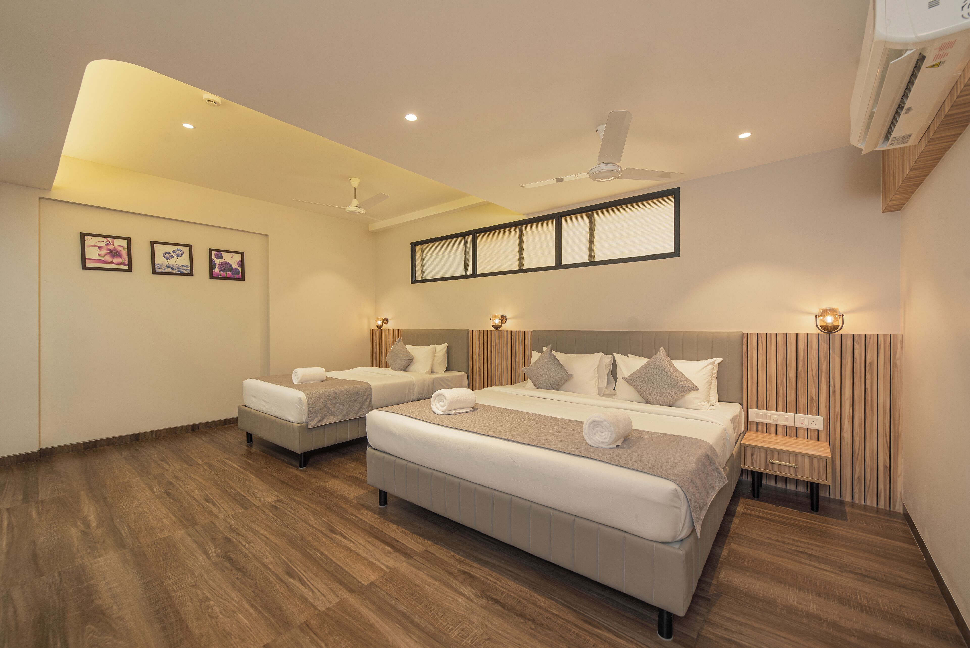 A modern hotel room with a large bed, two smaller beds, wooden flooring, ceiling fans, wall-mounted lights, and framed artwork.