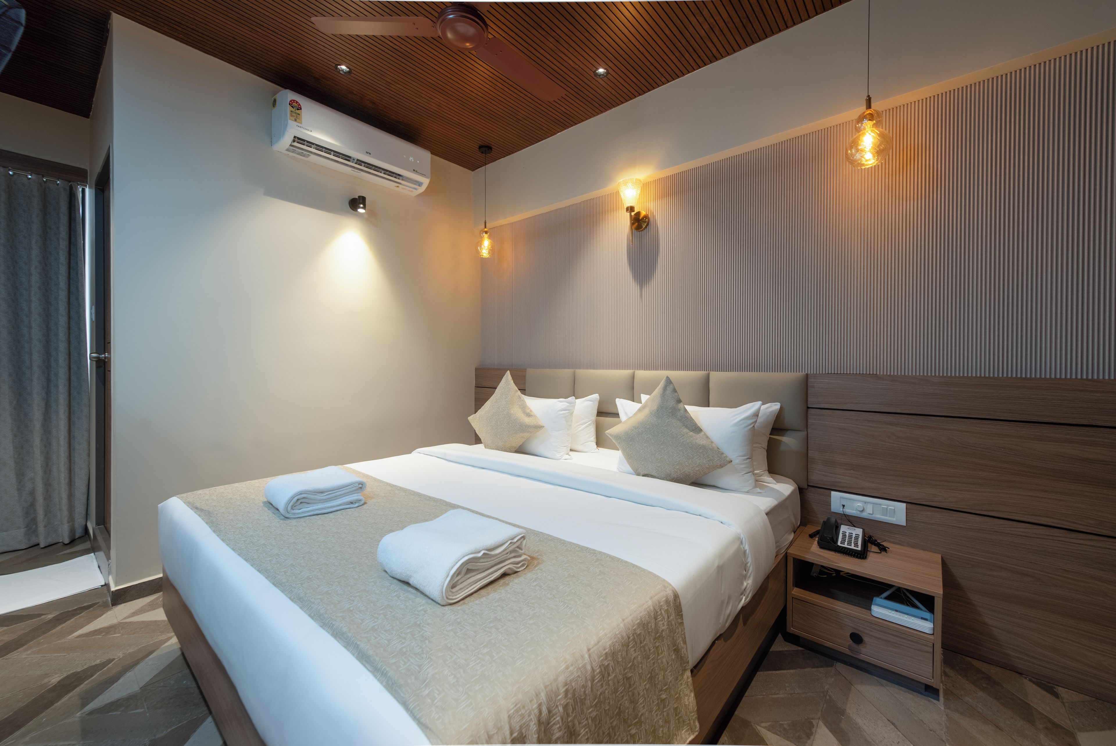 Quarto de hotel moderno com uma cama grande, mesa de cabeceira com telefone e controle remoto, e ar-condicionado fixado na parede.