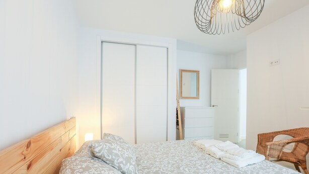 Ein Schlafzimmer mit einem Holz-Kopfteil, einer gemusterten Bettdecke, einem weißen Nachttisch, einem Korbstuhl und einem Spiegel an der Wand.