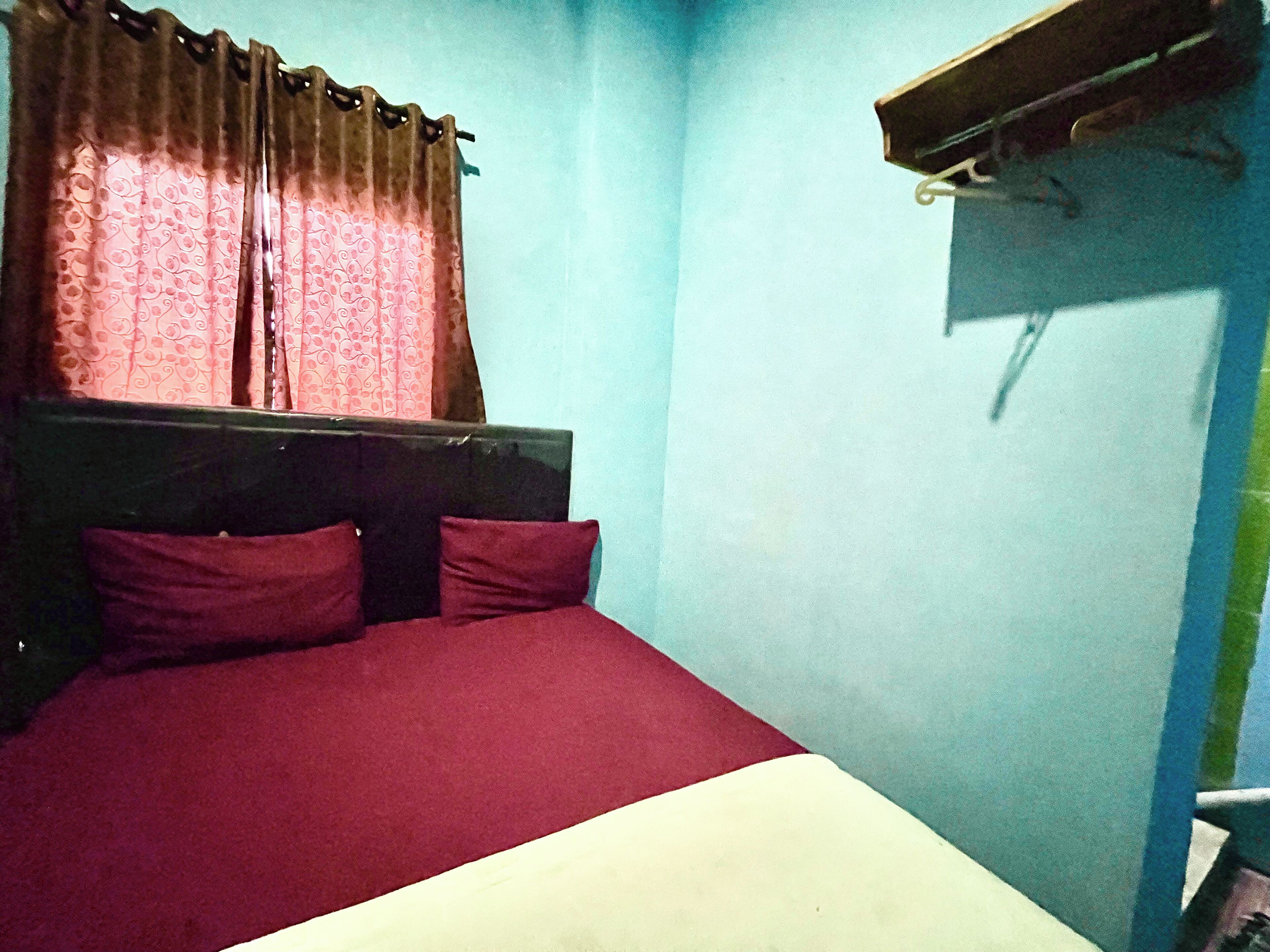 Ein kleines Zimmer mit einem Einzelbett, einem hölzernen Kopfteil, zwei Kissen und einer roten Bettdecke.