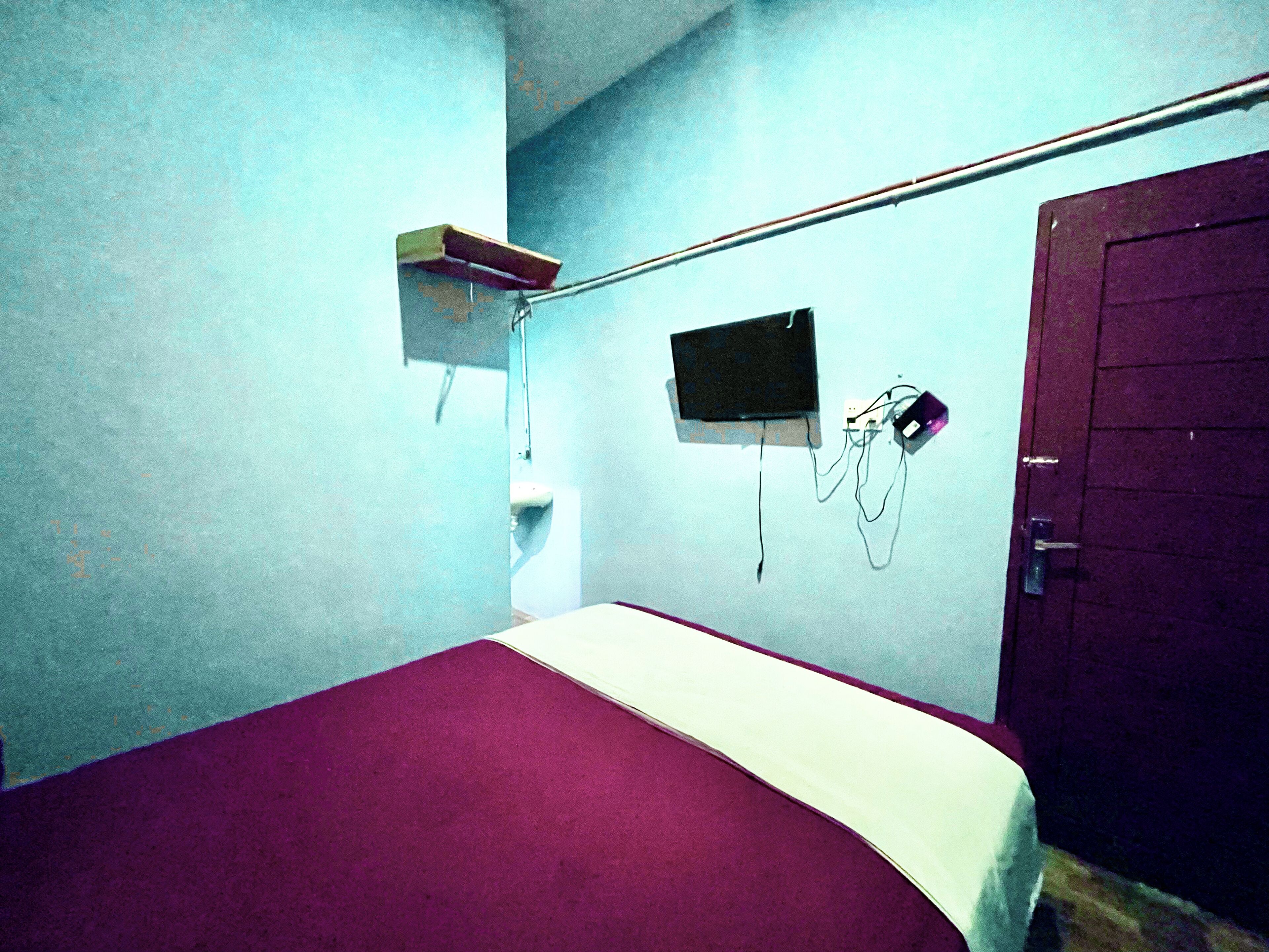 A small, sparsely furnished room with a bed, a television mounted on the wall, and a door.