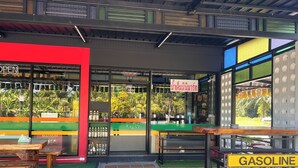 A storefront with a green and orange facade, a visible 'OPEN' sign, and a 'GASOLINE' sign outside. - Namkhem Inn 2 (Takua Pa)
