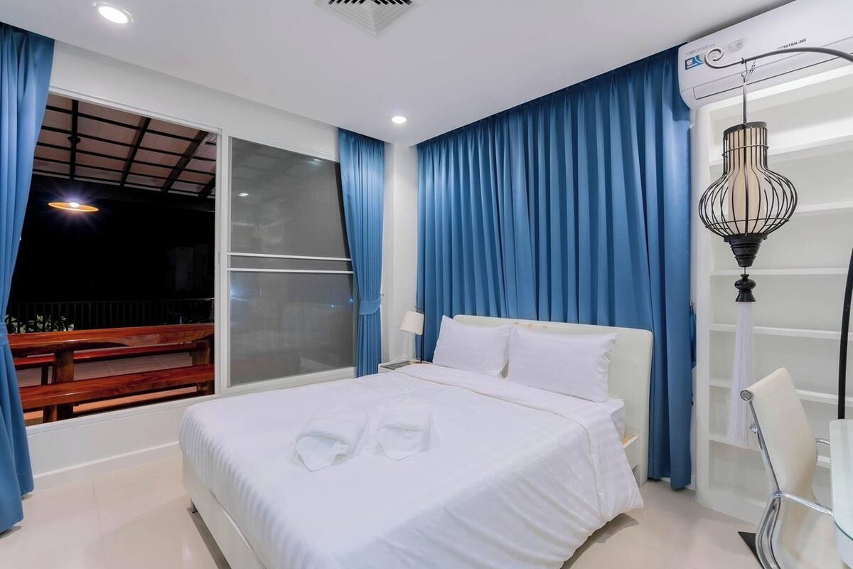 A hotel room with a large bed, a desk, and a chair. There is a window with blue curtains and a view of a balcony.