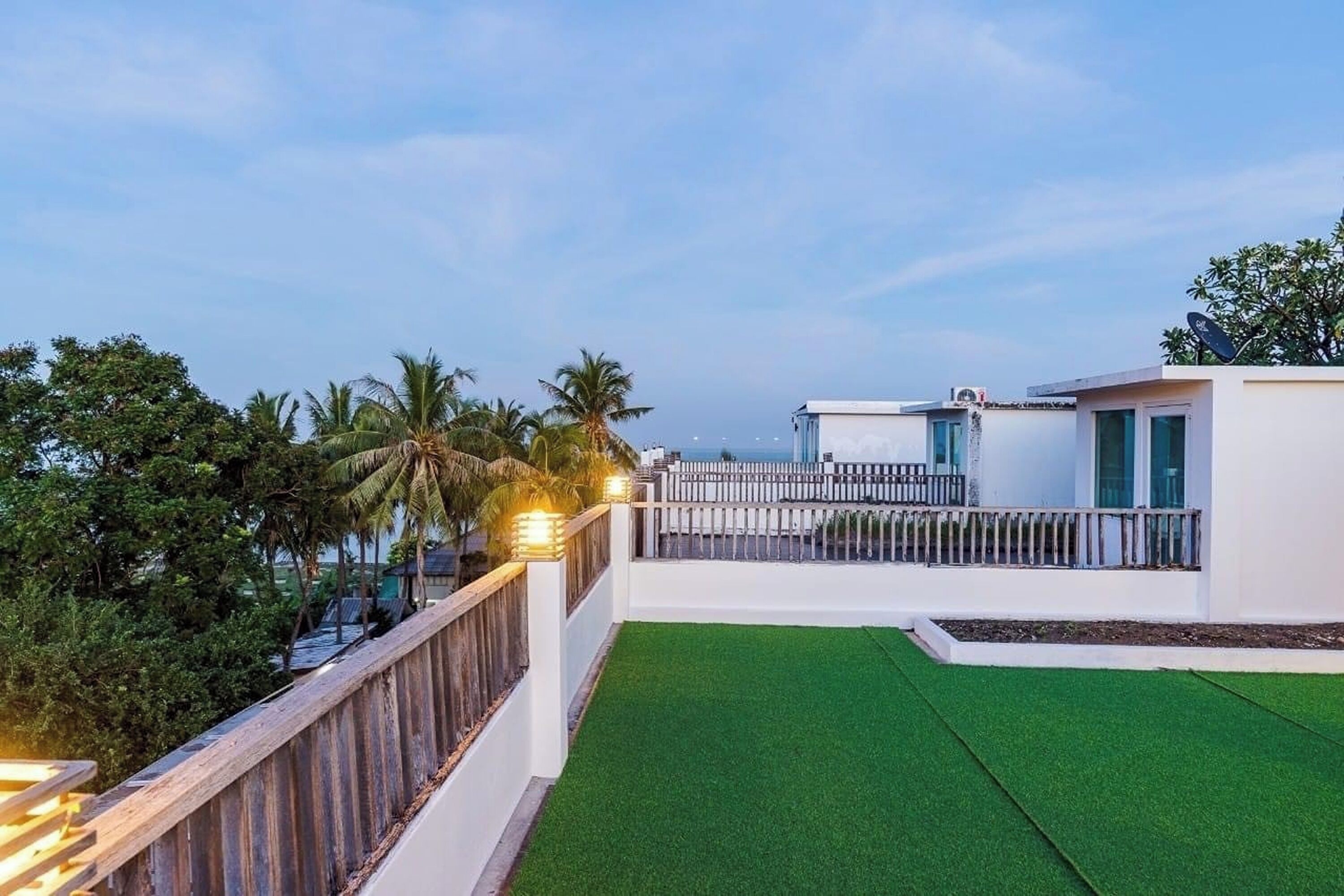 Une terrasse sur le toit avec un revêtement d’herbe artificielle, un bâtiment blanc doté d’un balcon, et des palmiers en arrière-plan.