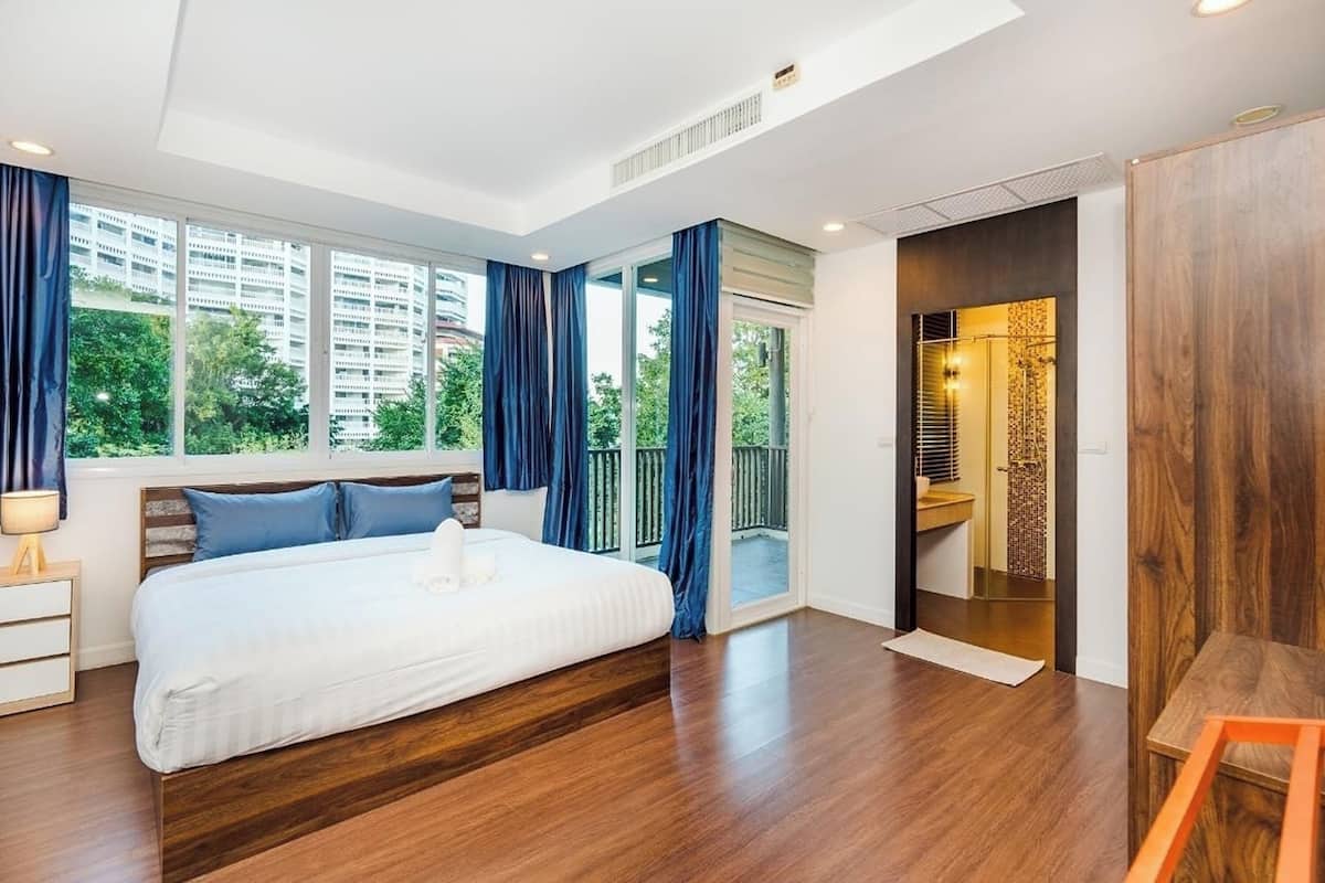 A modern bedroom with a large bed, wooden flooring, a view of greenery, and a bathroom with a shower and toilet.