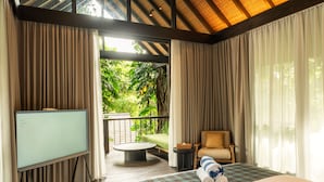 A hotel room with a bed, a TV, a balcony with a view of greenery, and a seating area with a chair and a table. - Rumah Lituhayu (Tabanan)