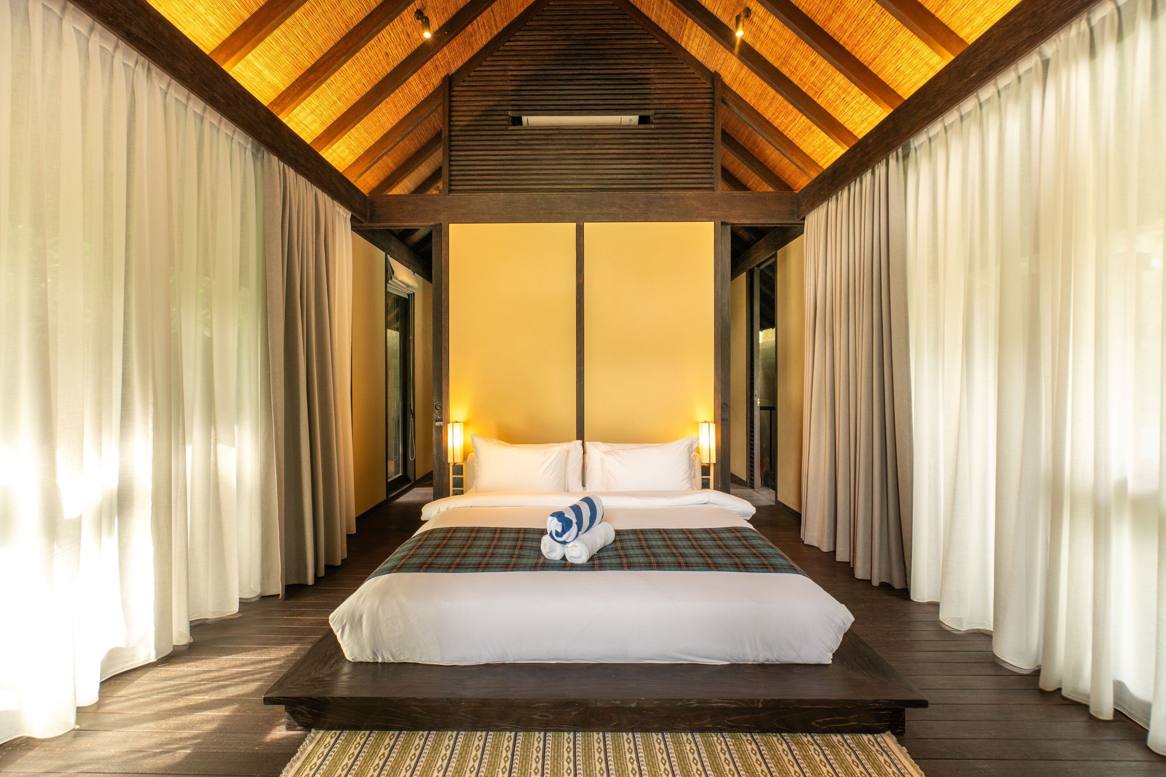 A large bed with white linens, a plaid blanket, and a rolled towel in a room with wooden beams and white curtains.