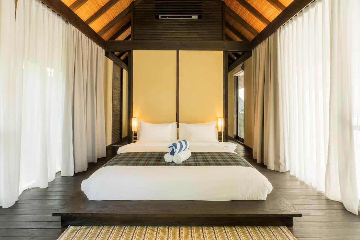 Una cama grande con sábanas blancas, una colcha escocesa y una toalla enrollada en el centro. La habitación tiene suelo de madera, techo de madera y cortinas blancas.