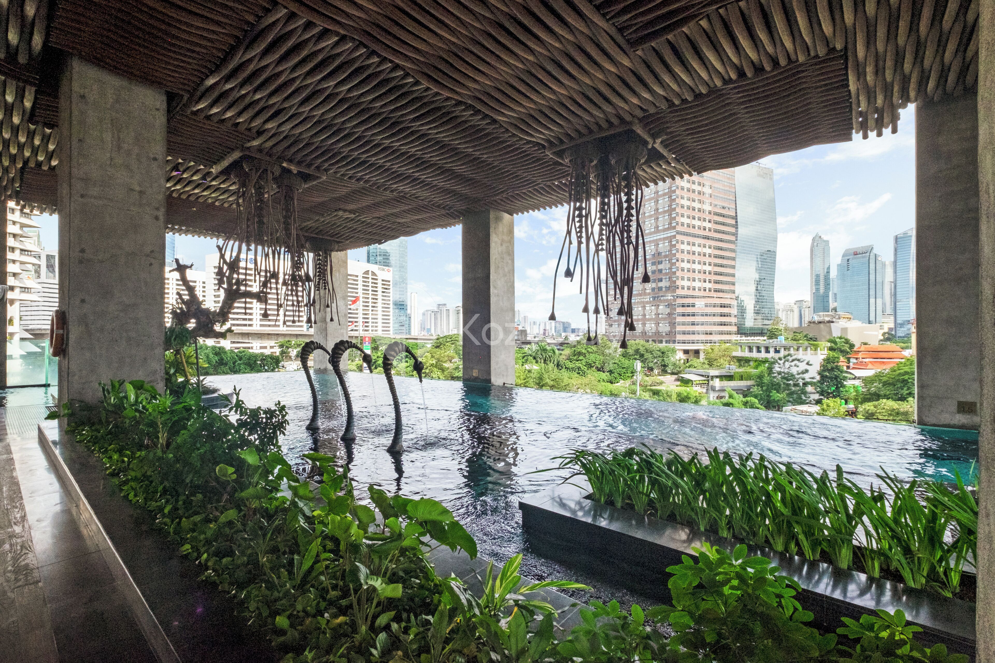 An infinity pool with a city view, surrounded by greenery and a unique wooden ceiling structure.