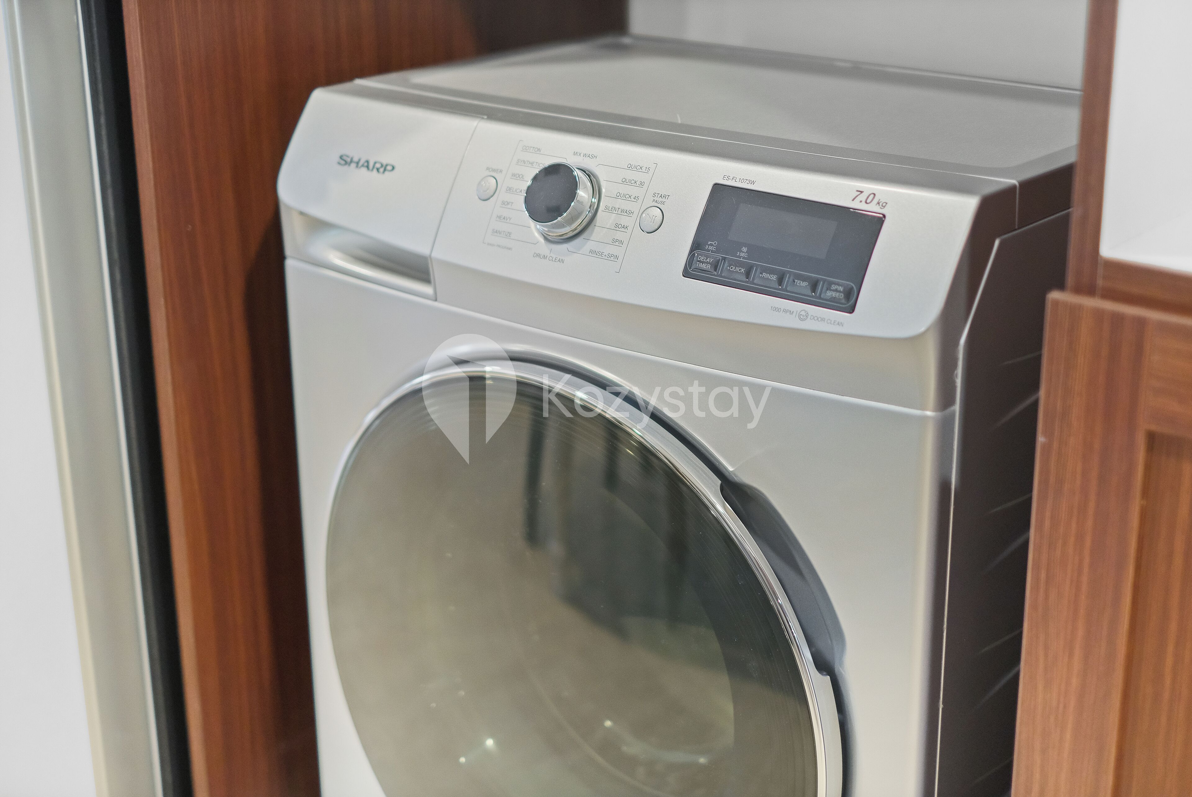 Vue de face d’un lave-linge moderne et compact, doté d’un écran numérique et d’un panneau de commande, le tout intégré dans un meuble en bois.
