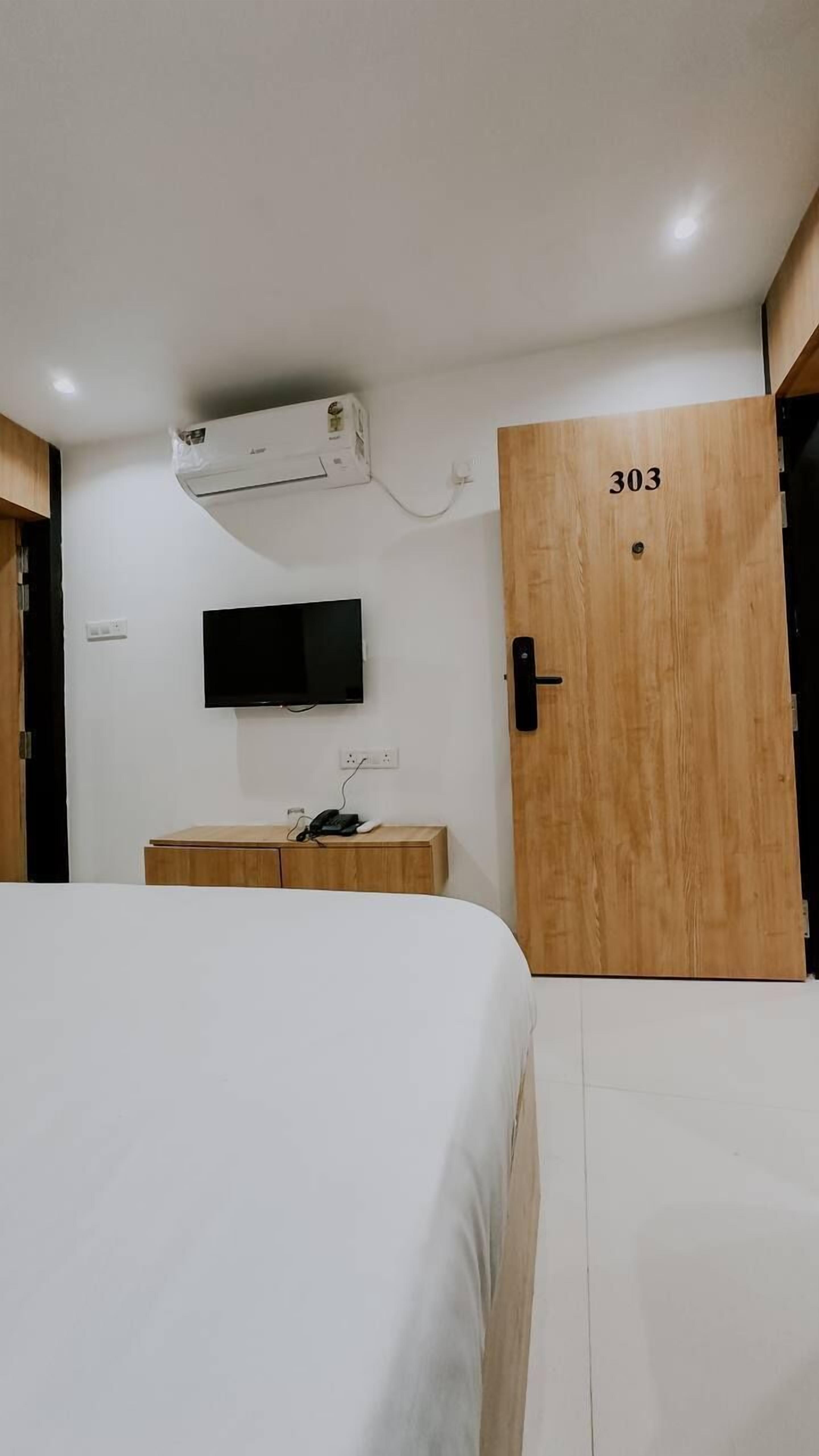 A hotel room with a bed, a TV mounted on the wall, an air conditioning unit, and a wooden door with the number 303.