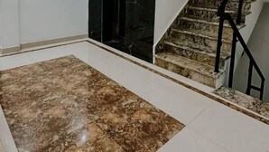 A spacious living area with a marble floor, a staircase with black railings, and a door leading to a dark room. - Nirvana Bliss (Prayagraj)