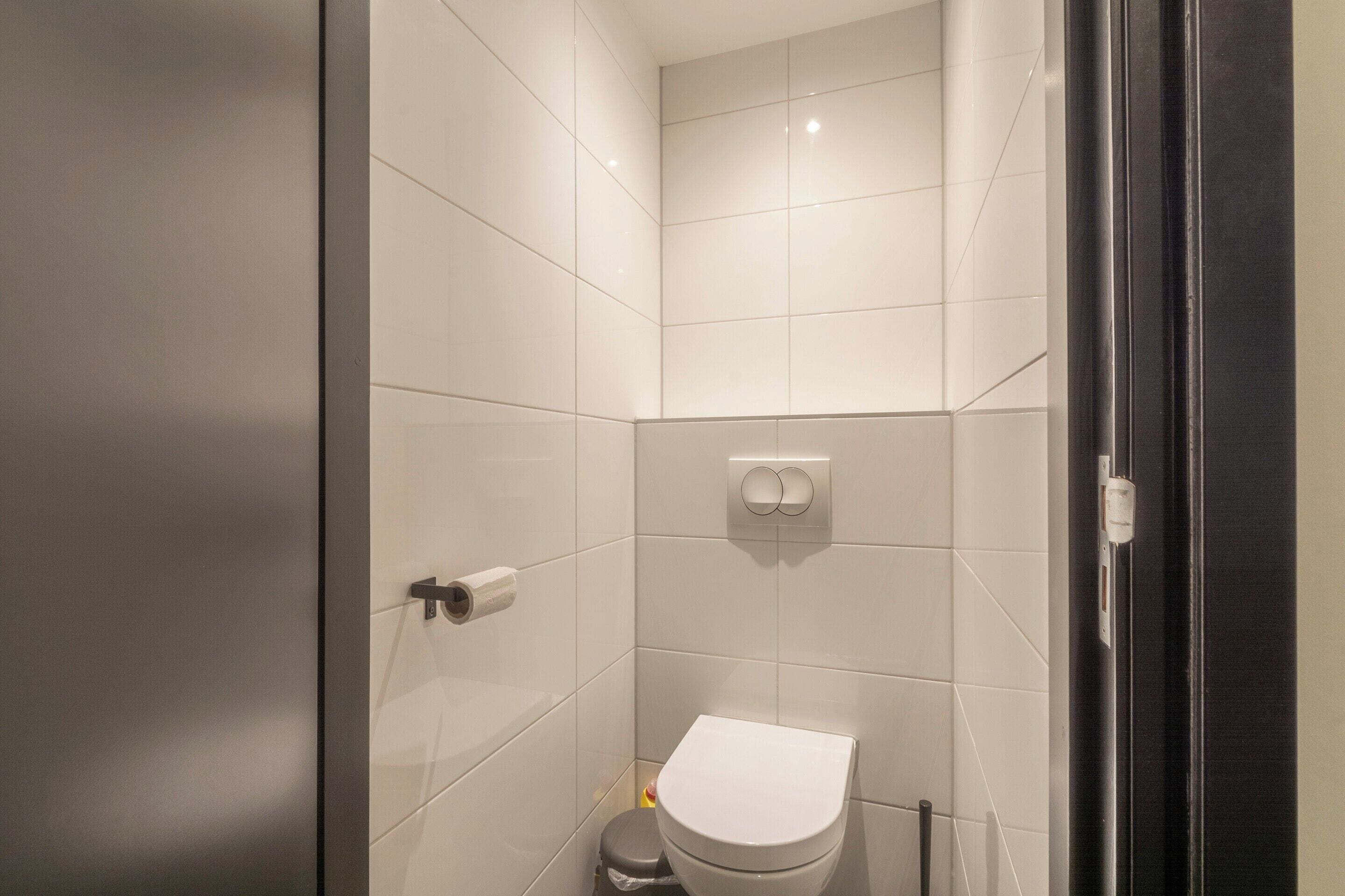 A small, modern bathroom with white tiles, a toilet, a trash bin, and a towel rack.