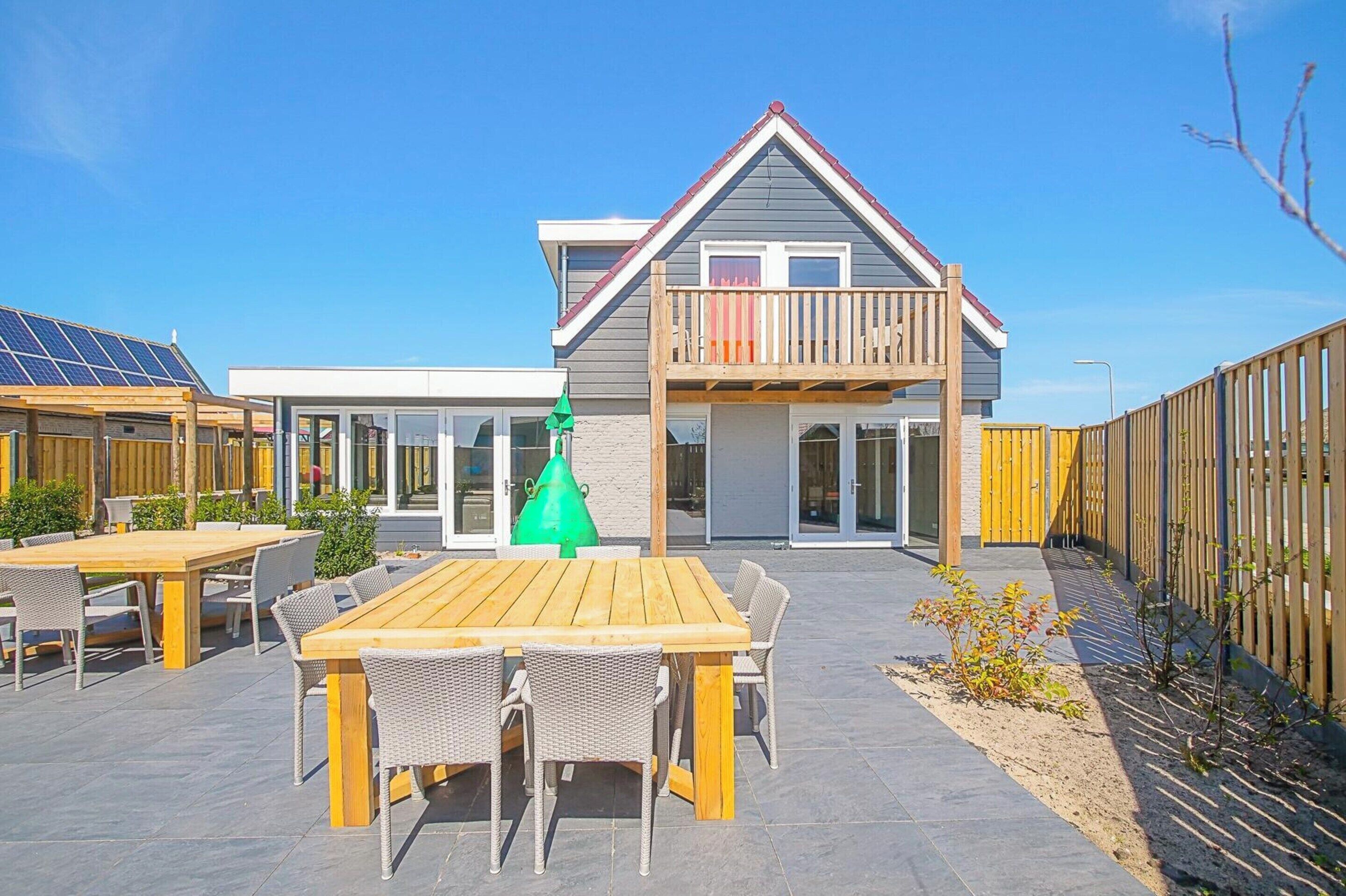 Una casa moderna con balcón, un patio con mesa y sillas de madera y un jardín cercado.