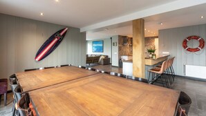 A spacious dining area with a long wooden table and multiple chairs. There is a surfboard mounted on the wall, and a lifebuoy is visible on the right side. - Callantsoog Group Getaway Beach (Callantsoog)