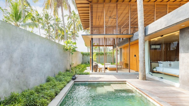 A modern outdoor pool area with a wooden deck, seating, and a view of a building with large windows. - Rumah Amadangi Kedungu (Tabanan)