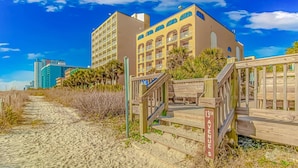 Een hotel aan het strand met een houten looppad naar de ingang, een helderblauwe lucht en een gebouw met meerdere balkons.