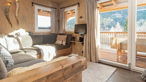 Interior acogedor de una cabaña con techo de madera, un sofá, un televisor y un balcón con vistas a las montañas.