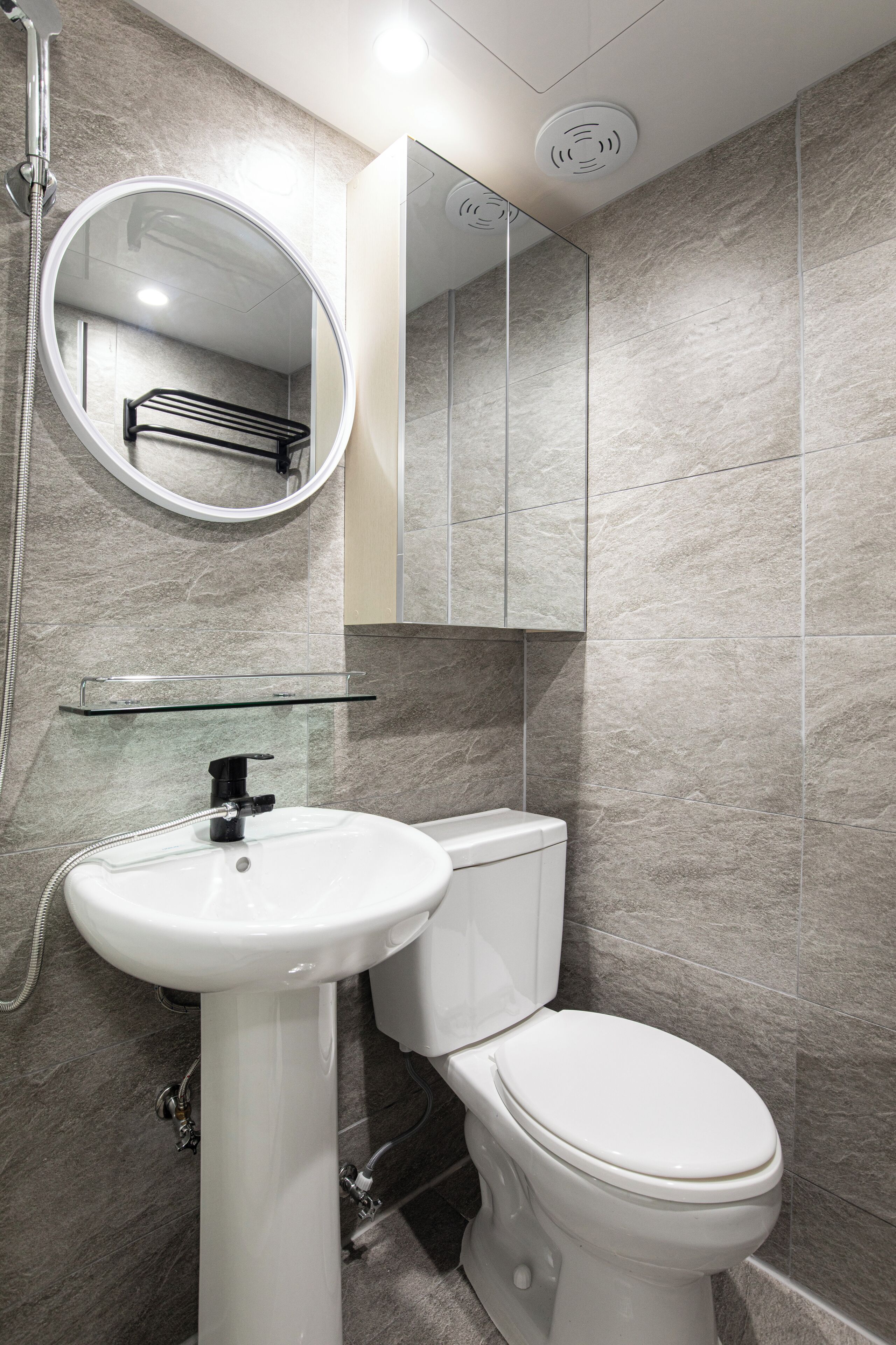 A modern bathroom with a white pedestal sink, a toilet, a round mirror, and a glass shelf above the sink.