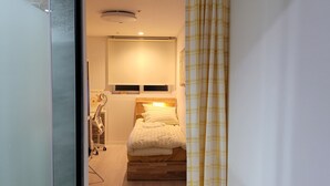 Quarto com cama, mesa de cabeceira e uma cadeira. Há uma janela com cortinas, uma luminária no teto e uma prateleira na parede.