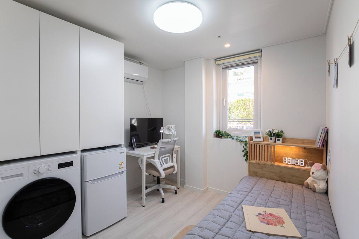 Habitación pequeña con lavadora, escritorio y silla, cama con colcha, ventana con vistas, y estante con libros y un oso de peluche.