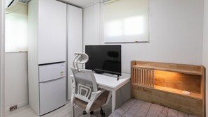 Quarto compacto com escrivaninha branca, monitor preto, cadeira branca, pequeno frigorífico branco e um banco de madeira com luz por baixo.