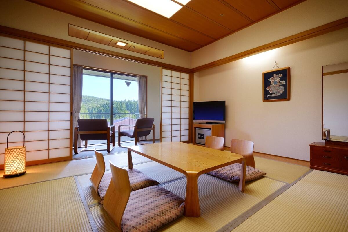 Et traditionelt japansk rum med tatamimåtter, et lavt bord, bløde siddepladser, et fjernsyn og en skydedør til en balkon.