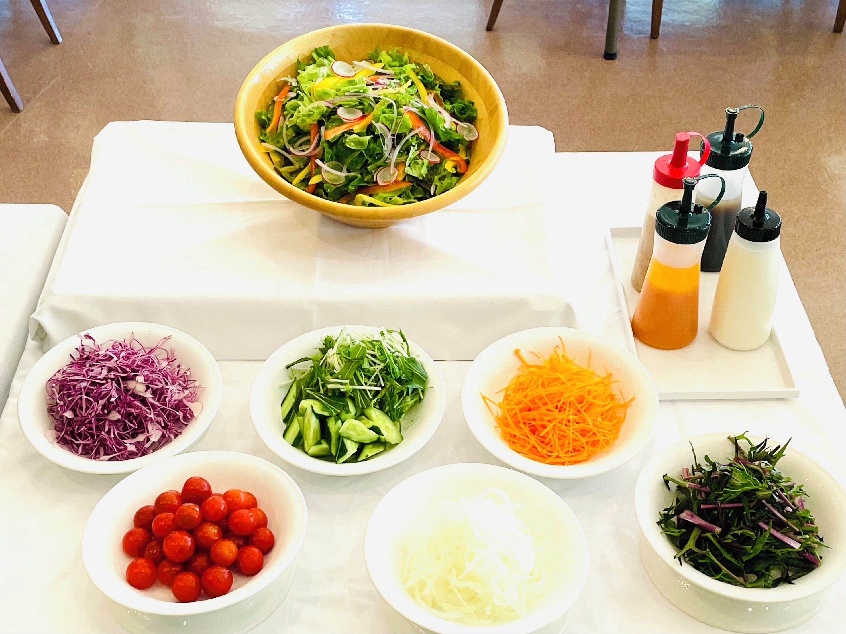 A table with various food items and condiments, including a bowl of salad, shredded vegetables, and small bowls of ingredients like tomatoes and cucumbers.