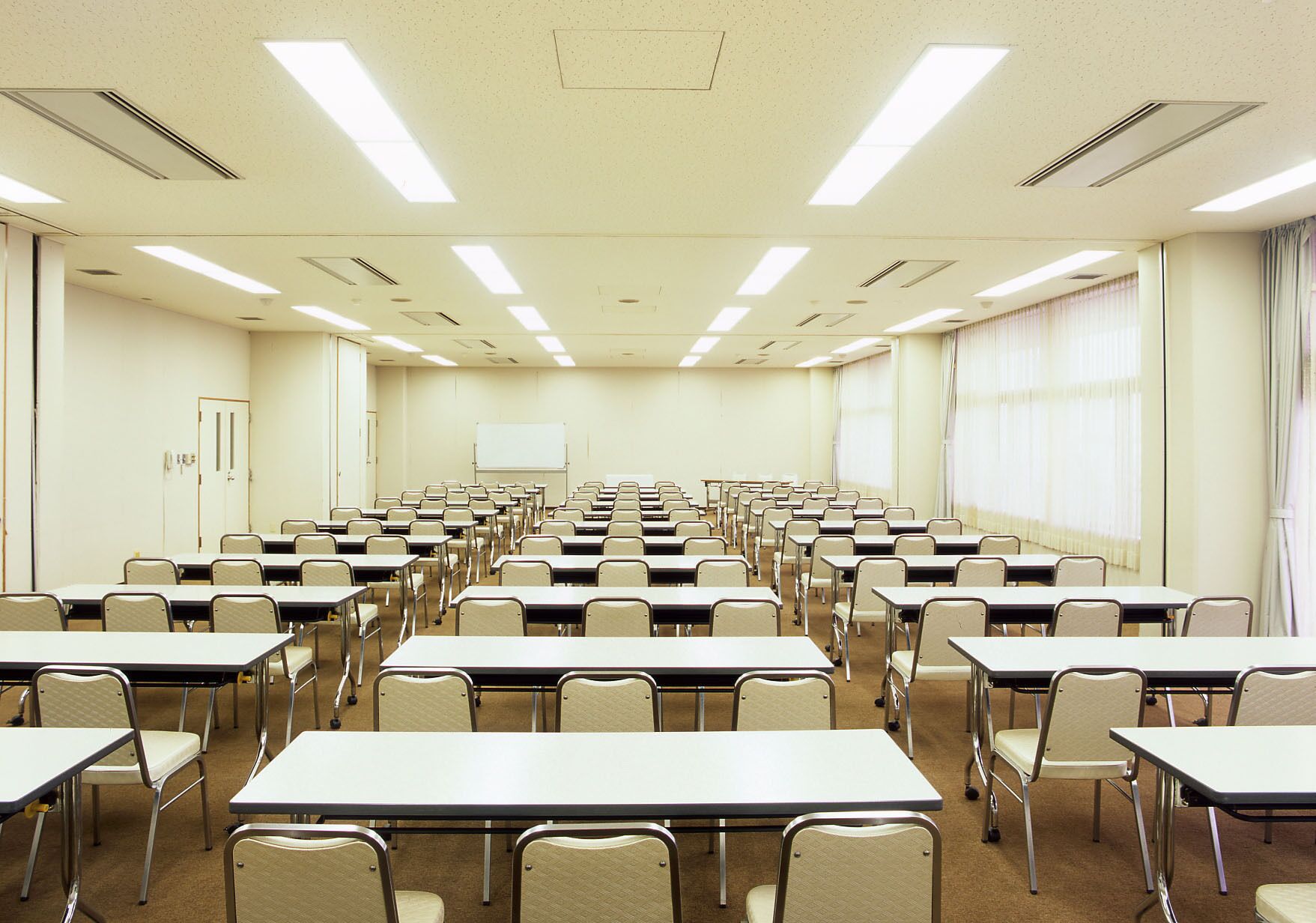 A conference room with rows of tables and chairs, a whiteboard, and large windows with curtains.