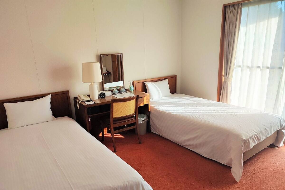 Et hotelværelse med to senge, et skrivebord med lampe, en stol, et lille bord og et spejl.