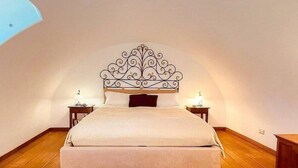Un dormitorio con techo curvo, una cama con cabecera decorativa, dos mesitas de noche con lámparas y un otomano azul.