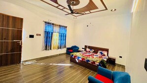 A bedroom with a wooden floor, a bed with a colorful quilt, a blue chair, and a ceiling fan. - The Corbett Canopy Home Stay (Kaladhungi)