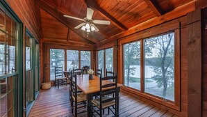 Une cabane en bois dotée d’un coin repas comprenant une table et des chaises, de grandes fenêtres et un ventilateur de plafond.