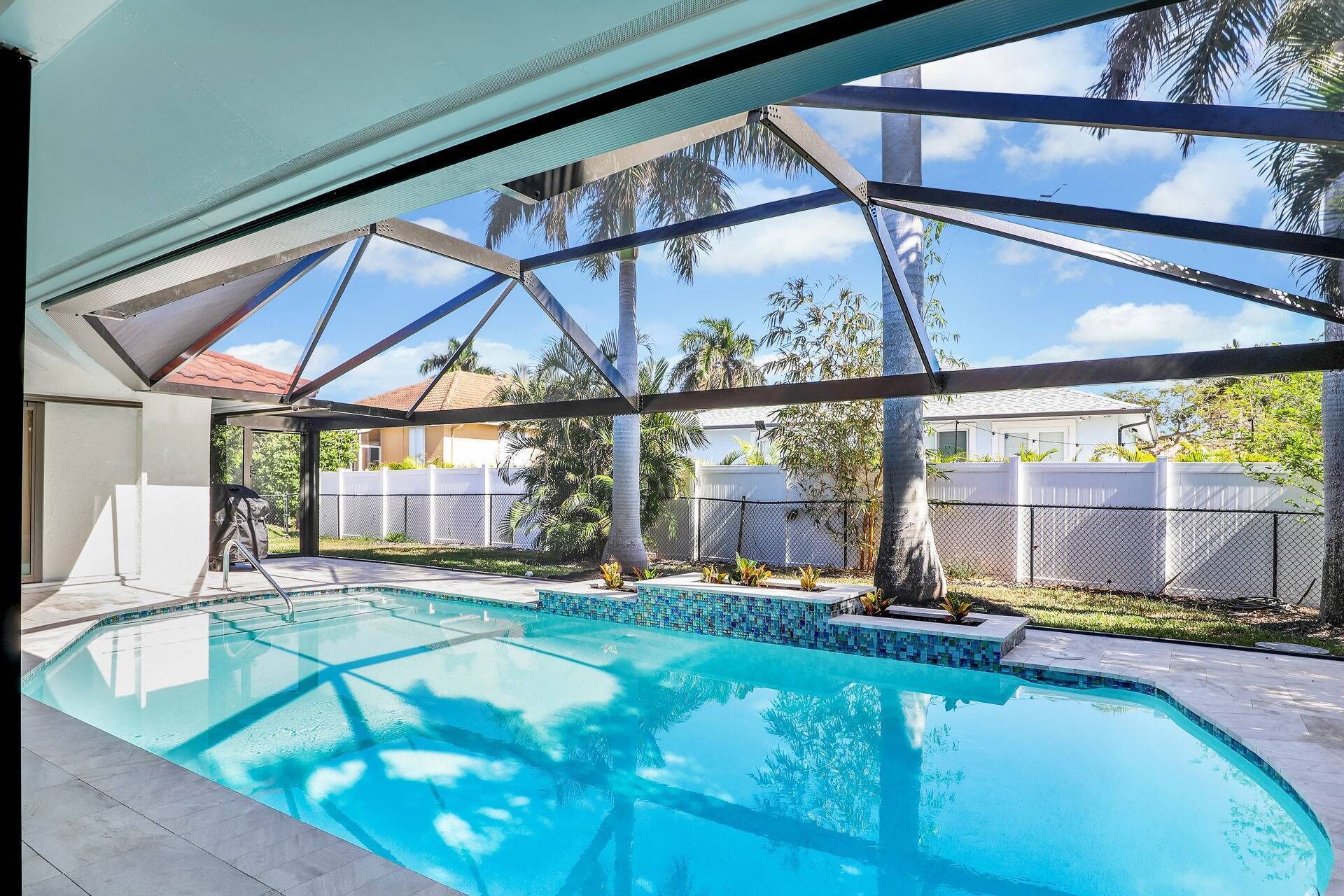 An indoor swimming pool with a glass ceiling, surrounded by a fenced area with palm trees and a view of neighboring houses.