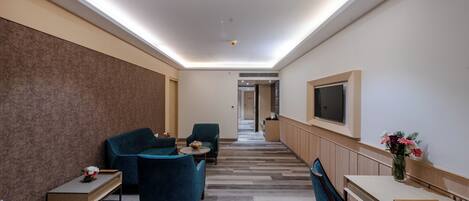 A modern hotel lobby with a long corridor, seating area with blue chairs, a wooden table, and a TV mounted on the wall.