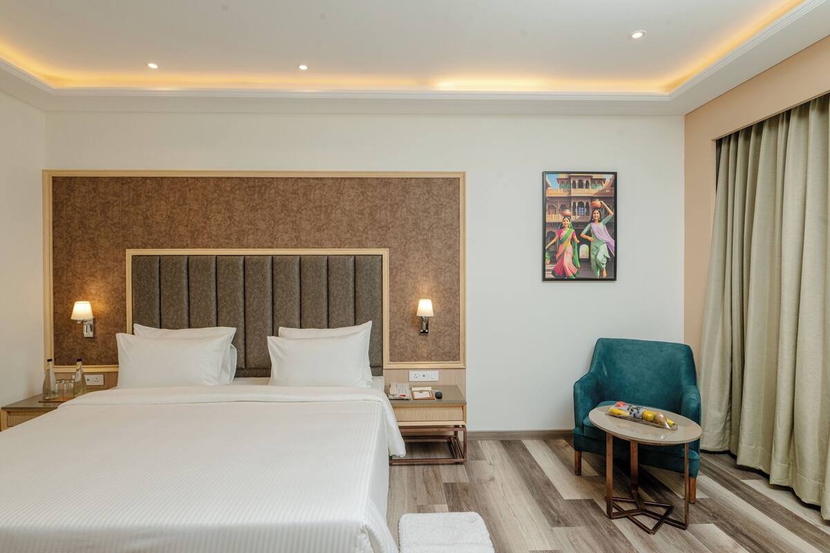 A hotel room with a large bed, a nightstand with a lamp, a small table with a chair, and a painting on the wall.