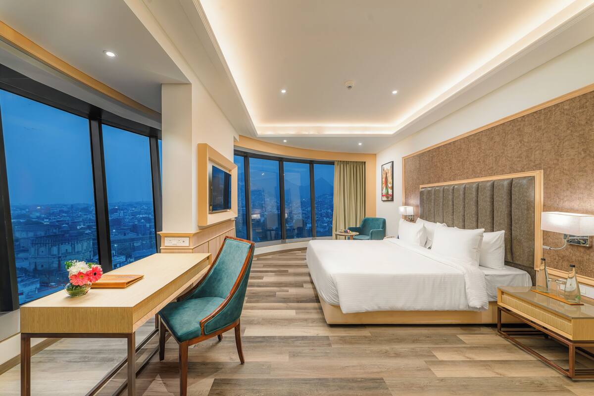 A hotel room with a large bed, a desk with a chair, a view of the city, and a large window.