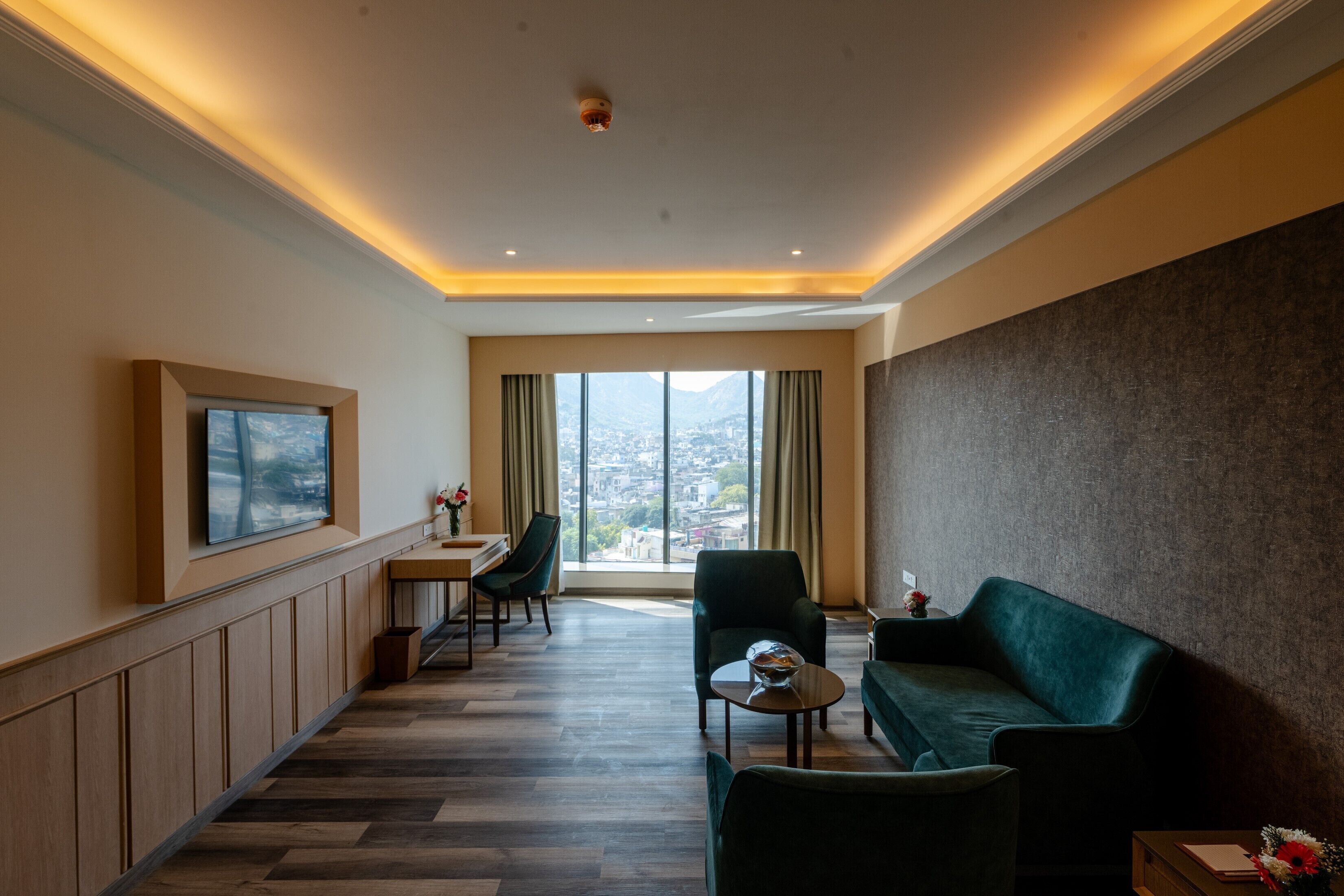 Een hotelkamer met een groot raam, een bank, een fauteuil, een klein tafeltje en een aan de muur bevestigde televisie.