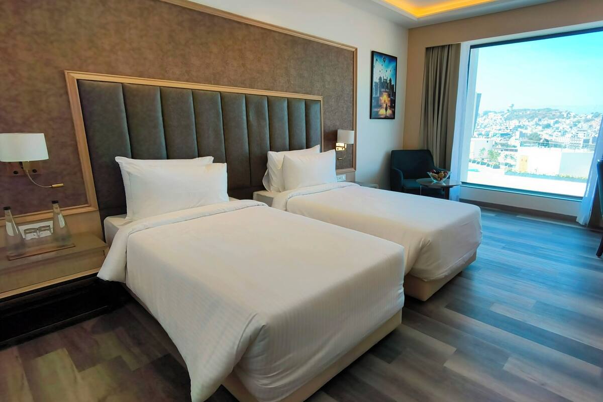 A hotel room with two beds, a desk, a chair, and a large window with a city view.