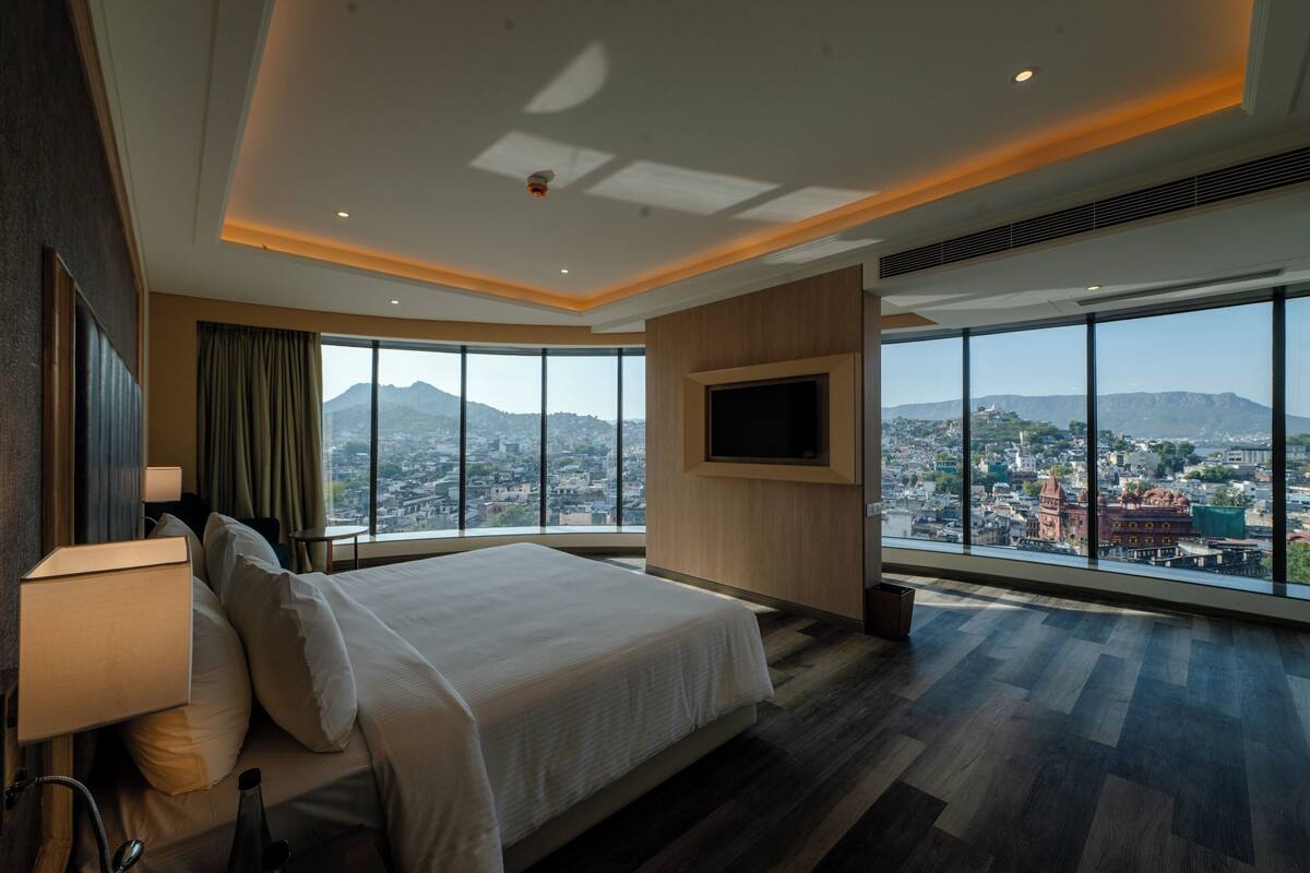 A modern hotel room with a large bed, a TV mounted on the wall, and a view of a cityscape through large windows.