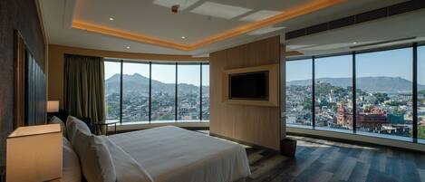 A modern hotel room with a large bed, a TV mounted on the wall, and a view of a cityscape through large windows.