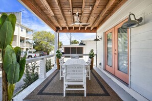 Een overdekt terras met een houten plafond, een witte tafel met stoelen, een potplant en een glazen schuindeur.